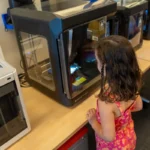 Female student awaits her creation during 3D printing in education.