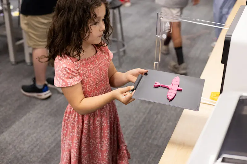  Little girl taking a tray with a pink 3D printed dragon out of a 3D printer.

