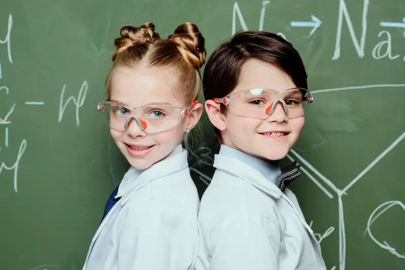 Smiling children wearing lab coats and protective glasses in front of a chalkboard.