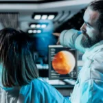 Two scientists stand in front of a control panel on a spaceship.