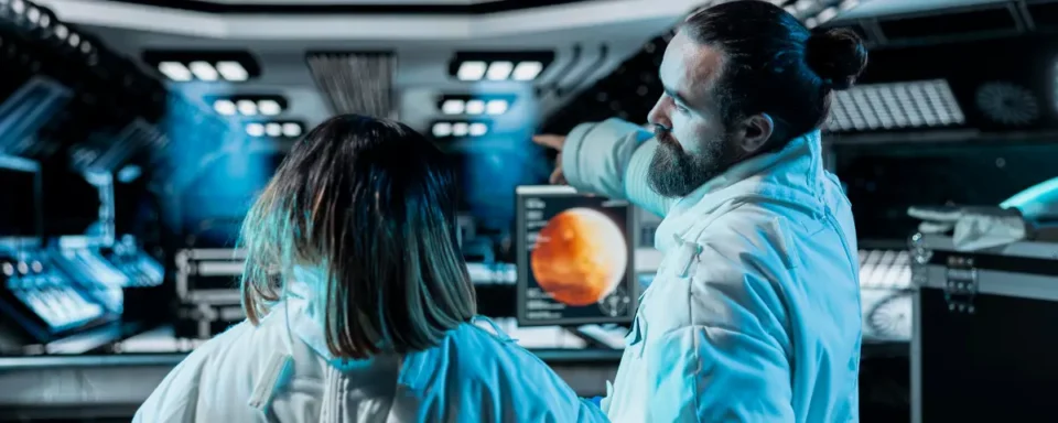 Two scientists stand in front of a control panel on a spaceship.