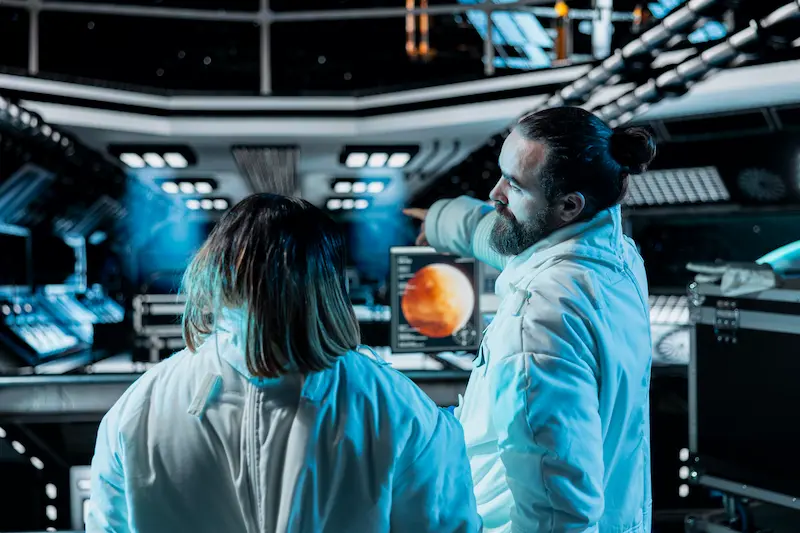 Two scientists stand in front of a control panel on a spaceship. 