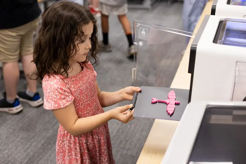 Girl taking a finished 3D printed design of a pink dragon out of a 3D printer.