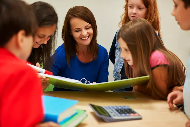 A smiling teacher showing several students sitting at a table a green binder.
