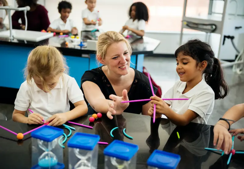 Group of young students working with blue and pink construction materials supervised by a teacher.
