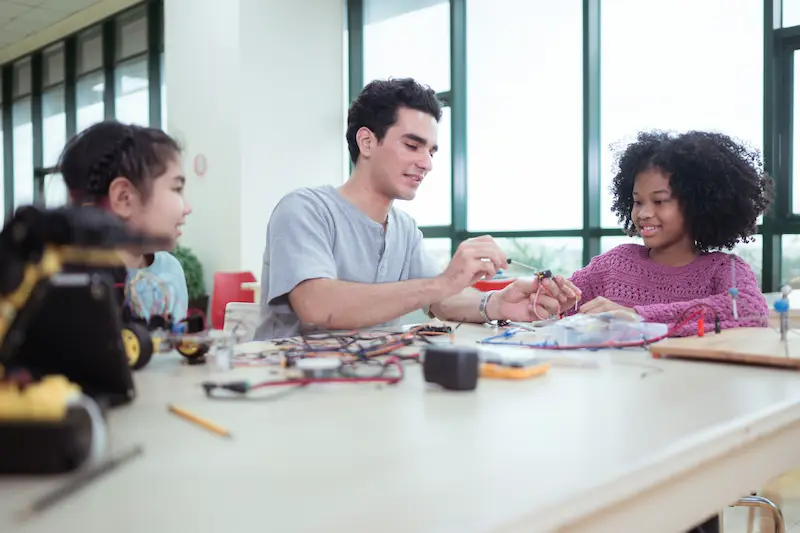 Teacher sitting at a table teaching a few students robotics.