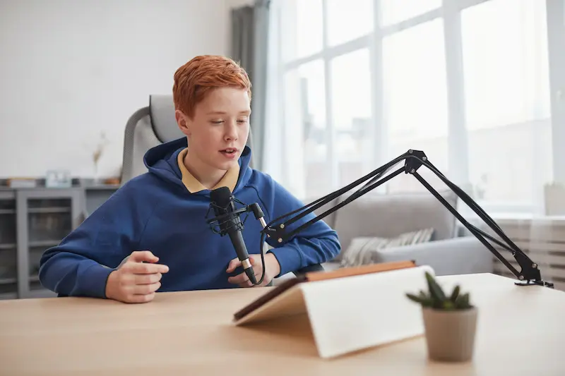 Teen with red hair sitting at a desk speaking into a podcasting microphone.
