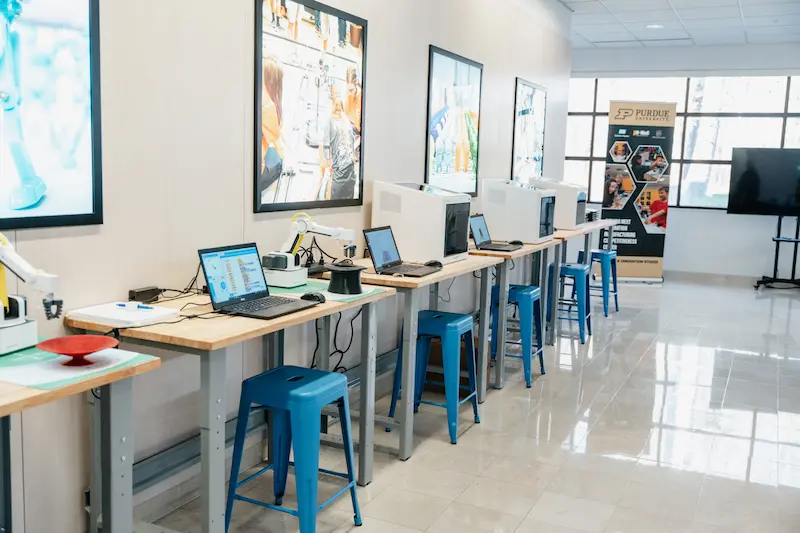 STEM lab with multiple stations, each with a blue chair, and a laptop. Some stations have robotic arms, and others have tabletop 3D printers. 
