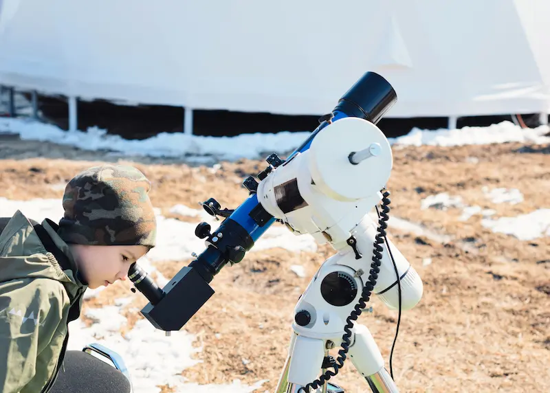 Boy looking into a blue and white telescope pointed at the sky.