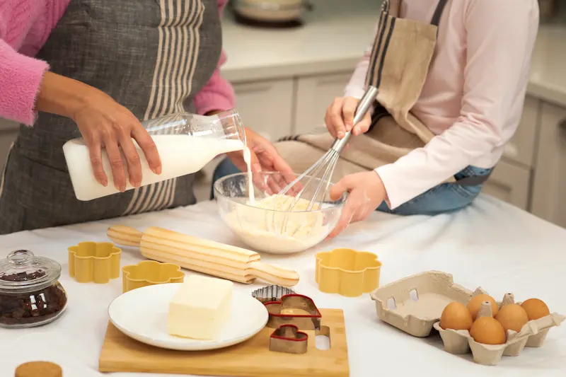 Child and adult baking together in a kitchen by mixing ingredients in a bowl.
