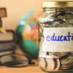 Glass jar full of coins labeled “education” in front of a stack of books and a pair of reading glasses.