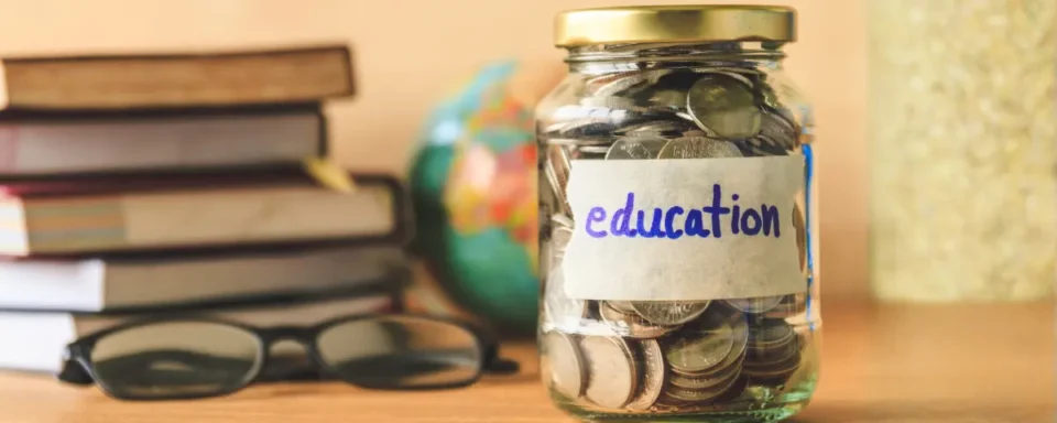 Glass jar full of coins labeled “education” in front of a stack of books and a pair of reading glasses.