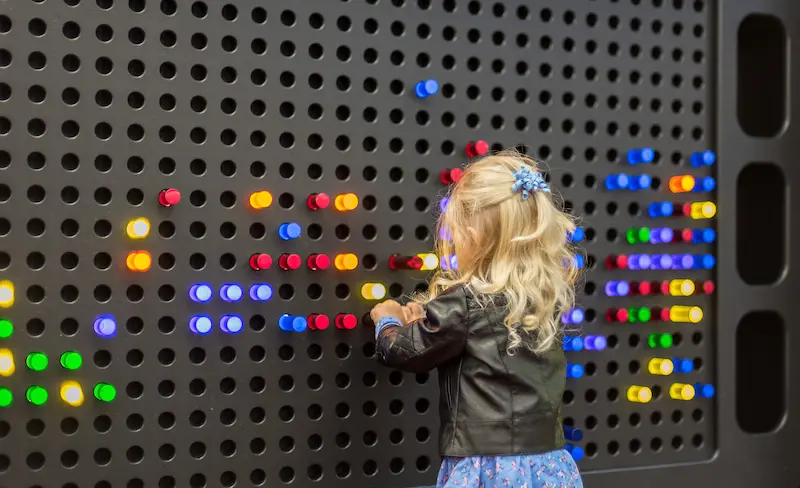A blonde-haired girl plays with an interactive light board with multiple colored lights, sorting them into colors.