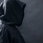 Person wearing a black cloak with a large hood in front of a grey background.