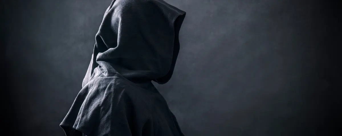 Person wearing a black cloak with a large hood in front of a grey background.