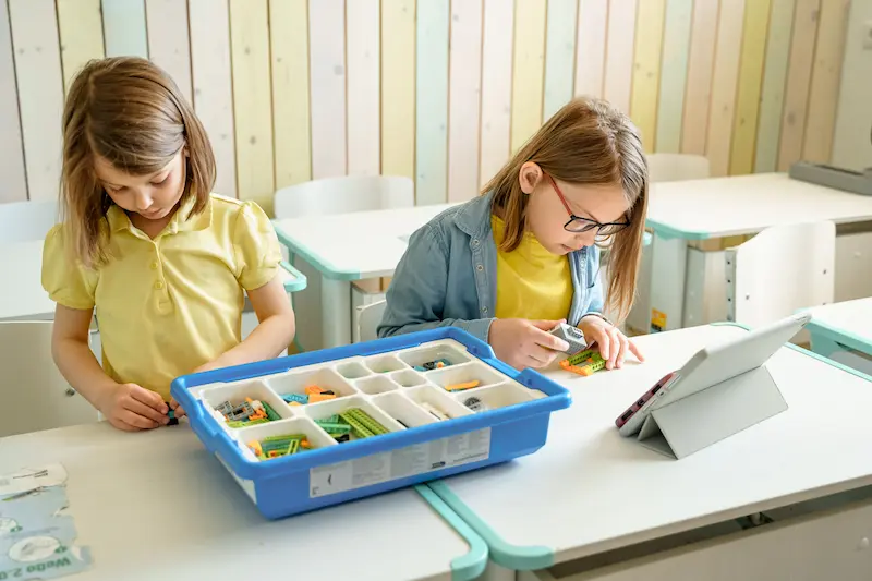 Two young girls are working on a project using materials from a well-organized bin.