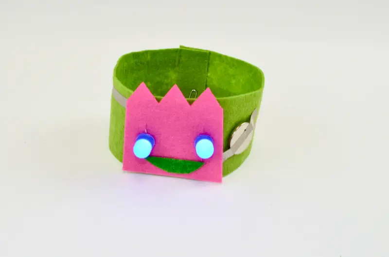 Green fabric bracelet with a pink face sewn on and two glowing blue eyes.