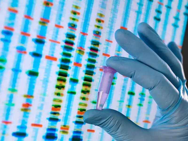 A gloved hand holding a tube of liquid in front of a screen displaying a gene sequence.