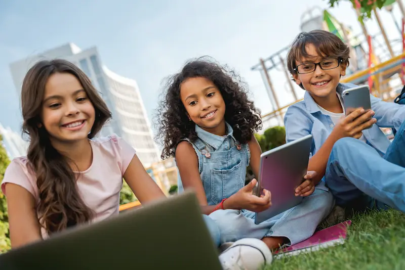 Three smiling children sitting on the grass, each using a tablet or a laptop.