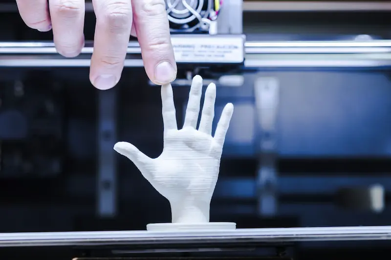 A white 3D-printed model of a hand. A human hand is touching the fingertip of the smaller model.