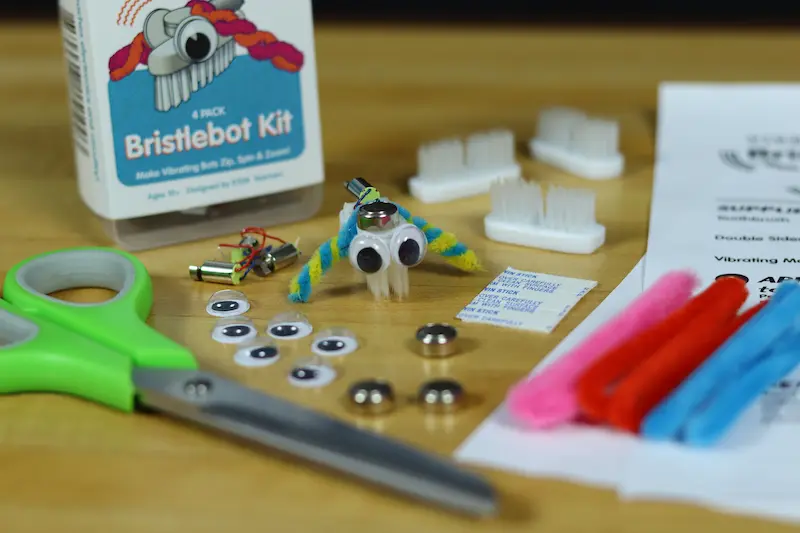 A completed Bristlebot with googly eyes and yellow and blue legs made out of pipe cleaners. There are more supplies, like scissors and more pipe cleaners, in the background.