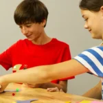 Two students assembling Brown Dog Gadgets kits at a table with craft supplies, like colored pencils, paper, and scissors.