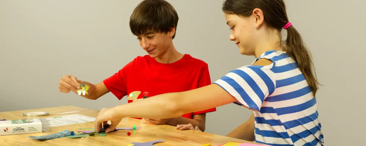 Two students assembling Brown Dog Gadgets kits at a table with craft supplies, like colored pencils, paper, and scissors.