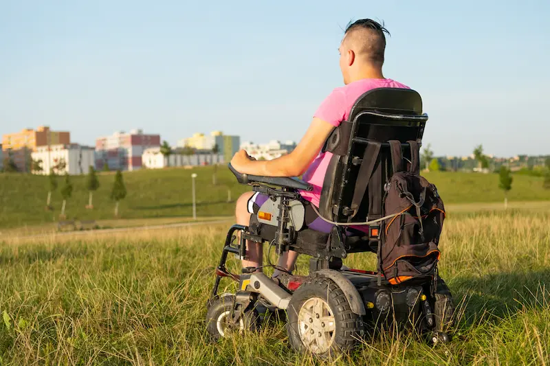 Young man in a motorized wheelchair in a grassy park.
