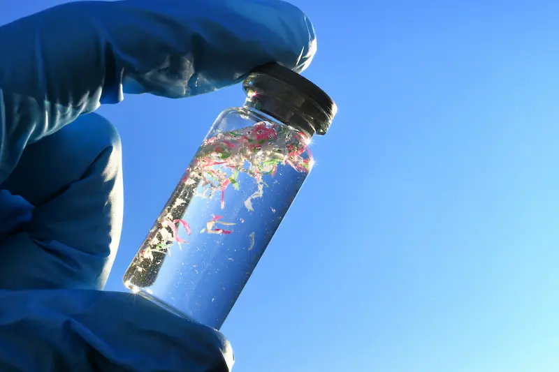 Gloved hand holding a glass vial filled with microplastics floating in water.
