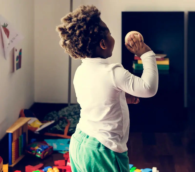 Small curly-haired child throwing a baseball indoors.
