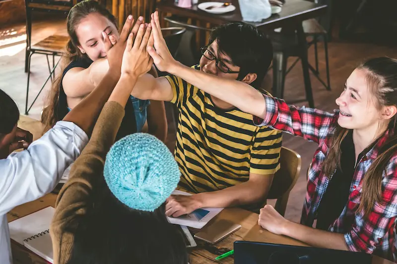 Group of students sitting around a table in a classroom sharing a group high-five.