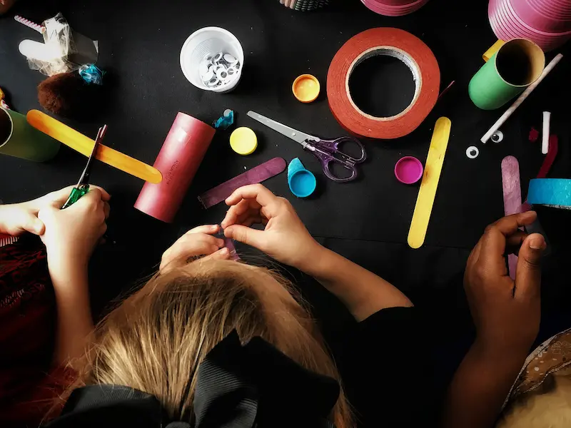Children doing arts and crafts projects at a black table with colorful paper, tape and other craft supplies.