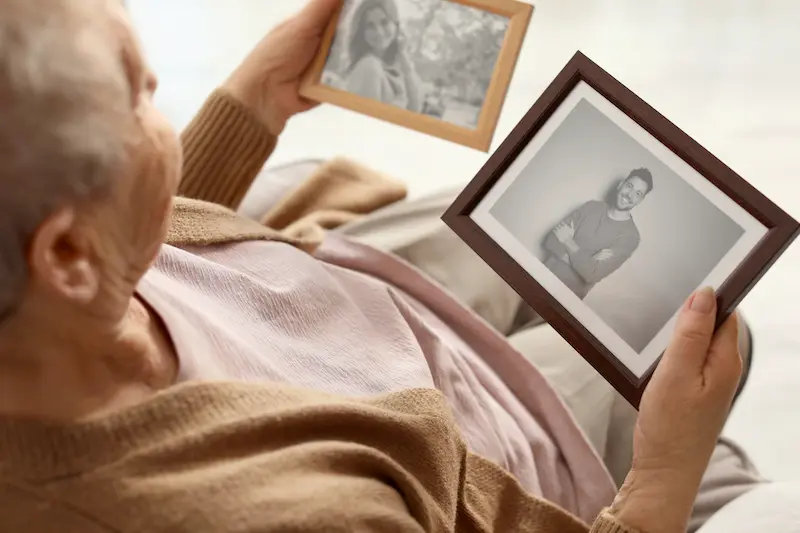 Old woman looking at black and white framed photographs indoors.