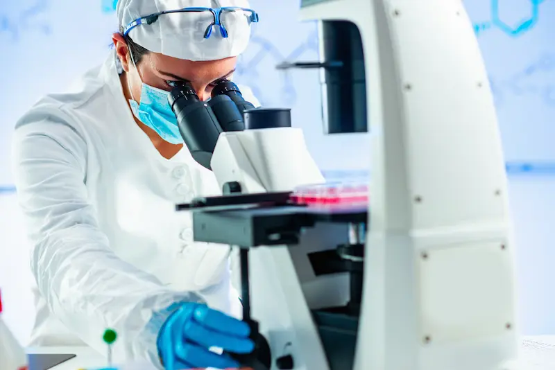 Scientist wearing a lab coat and gloves looking through a microscope.